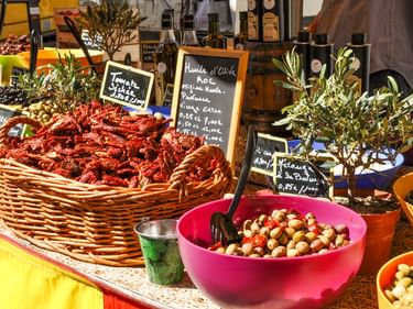 Marktstand in der Provence mit getrockneten Tomaten im Weidenkorb, Oliven in rosa Schüssel, Olivenölflaschen und kleinen Olivenbäumen mit Kreidetafeln.