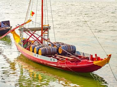 Rot-gelbes traditionelles Holzboot mit Weinfässern auf ruhigem Wasser. Das dekorierte Schiff hat einen Mast und eine bunte portugiesische Flagge.