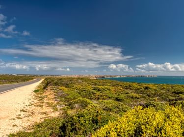Asphaltierte Straße entlang der portugiesischen Küste bei Sagres mit niedriger Vegetation, türkisfarbenem Meer und dramatischen Wolken.