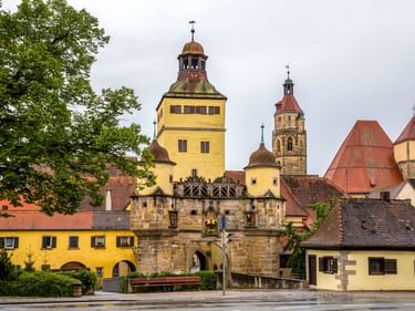 Mittelalterliches Ellinger Tor mit gelbem Turm und Steinbogen in Weißenburg. Kirchtürme und traditionelle Gebäude dahinter unter bewölktem Himmel.