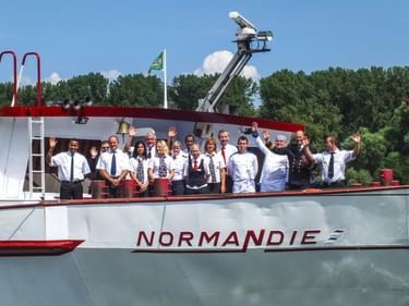 Besatzungsmitglieder in weißen Uniformen stehen auf dem Oberdeck des Schiffes Normandie und winken in die Kamera, mit Bäumen und blauem Himmel im Hintergrund.