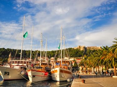 Drei hölzerne Segelboote mit hohen Masten liegen im Hafen von Hvar in Kroatien. Palmen säumen die Uferpromenade mit historischen Gebäuden am Hang.