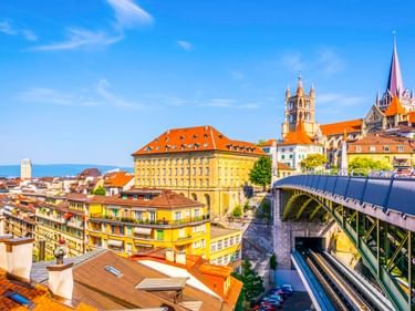 Panoramablick auf Lausanne mit bunten Gebäuden, einer modernen Brücke und der Kathedrale Notre-Dame mit ihrem markanten Turm vor blauem Himmel.