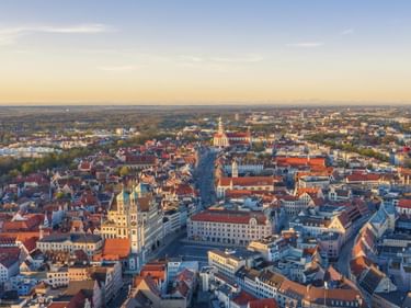 Luftaufnahme der Augsburger Altstadt bei Sonnenuntergang mit historischen Gebäuden, roten Ziegeldächern und Kirchtürmen. Die Stadtlandschaft erstreckt sich bis zum Horizont.