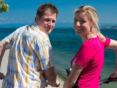 Mann im karierten Hemd und Frau in pinkem Oberteil mit Fahrrädern am Bodensee. Blaues Wasser und klarer Himmel im Hintergrund.