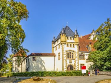 Burg Jagsthausen mit gelbem Turm, grauen Kegeldächern und weißen Mauern, umgeben von grünen Bäumen unter blauem Himmel. Besucher am Eingang.