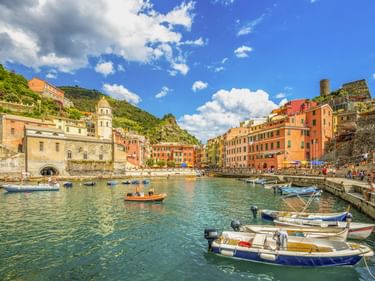 Hafen von Vernazza mit Fischerbooten im türkisfarbenen Wasser, umgeben von bunten Häusern an Hängen unter blauem Himmel mit weißen Wolken.