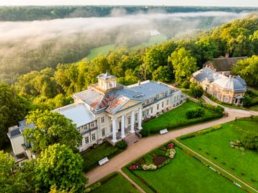 Luftaufnahme von Krimuldas Manor in Sigulda mit formalen Gärten und Blumenbeeten, umgeben von dichtem Wald mit Morgennebel im Tal.