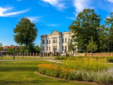 Weiße neoklassizistische Villa mit verzierter Fassade, umgeben von Parkanlage mit Blumenbeeten, grünem Rasen und alten Bäumen unter blauem Himmel.