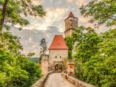 Steinbrücke zur Burg Zvíkov mit rotem Ziegeldachturm und rundem Turm, eingerahmt von grünen Bäumen unter bewölktem Himmel.