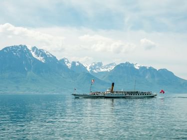 Historischer Raddampfer mit Schweizer Flagge auf dem Genfersee mit schneebedeckten Alpen im Hintergrund unter bewölktem Himmel.