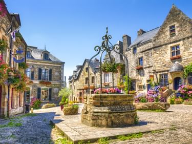 Kopfsteinpflasterplatz in Rochefort mit Steinbrunnen, traditionellen französischen Gebäuden mit blauen Fensterläden und bunten Blumen.