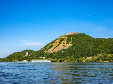 Burg Visegrad auf einem grünen bewaldeten Hügel mit Blick auf die Donau. Ein Passagierschiff fährt auf dem blauen Wasser unter klarem Himmel.