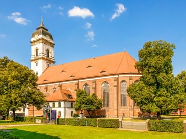 Dom St. Marien in Fürstenwalde mit orangerotem Dach, weißem Glockenturm und Backsteinmauern, umgeben von grünen Rasenflächen und Bäumen.