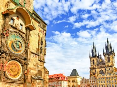 Prager Astronomische Uhr am Altstädter Rathausturm mit bunten Zifferblättern, Teynkirche im Hintergrund unter blauem Himmel.