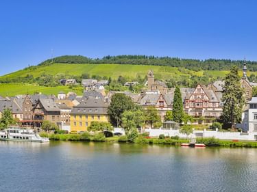 Traben-Trarbach an der Mosel mit Fachwerkhäusern, einem Passagierschiff und weinbergbedeckten Hügeln unter blauem Himmel.