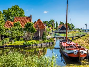Rote Fischerhäuschen entlang eines Kanals in Enkhuizen mit einem braunen Boot. Grüne Bäume und eine Brücke unter blauem Himmel.