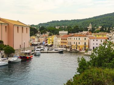 Bunter Hafenort Lošinj mit Booten im Wasser, mehrfarbigen Gebäuden entlang der Uferpromenade und grünen Hügeln im Hintergrund.