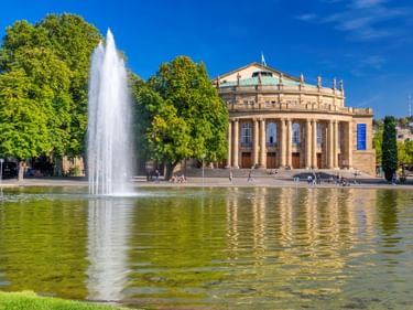 Stuttgarter Staatstheater, ein neoklassizistisches Gebäude mit Säulen, spiegelt sich in einem Teich mit hohem Springbrunnen. Grüne Bäume unter blauem Himmel.