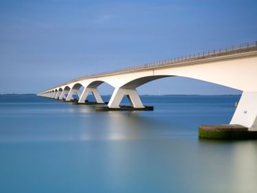 Lange weiße Brücke mit markanten Bogenpfeilern über ruhigem blauen Wasser unter klarem Himmel. Die Zeelandbrücke zeigt sich mit Bögen.
