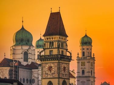 Passauer Skyline bei Sonnenuntergang mit Stadtpfarrkirche St. Paul mit grünen Zwiebeltürmen, Uhrturm und Dom St. Stephan vor orangefarbenem Himmel.