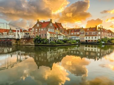 Historische Gebäude am Wasser in Enkhuizen mit roten Ziegeldächern spiegeln sich bei Sonnenuntergang in ruhigem Hafenwasser.