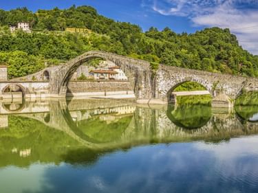 Ponte del Diavolo, eine mittelalterliche Steinbrücke mit mehreren Bögen, die sich im ruhigen Flusswasser spiegelt, umgeben von grünen Hügeln in Italien.