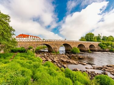 Historische Steinbogenbrücke Tullbron über einen Fluss in Falkenberg mit vier Bögen, grüner Vegetation an den Ufern und weißem Gebäude mit rotem Dach.
