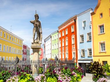 Sankt-Georgs-Brunnen in Schärding mit bunten Barockgebäuden in Gelb, Orange, Grün und Blau dahinter. Blumenbeete umgeben das Denkmal.