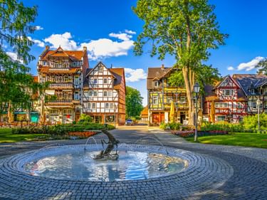 Runder Brunnen auf gepflastertem Platz, umgeben von bunten Fachwerkhäusern in Bad Sooden-Allendorf. Grüne Bäume rahmen die Szene unter blauem Himmel.