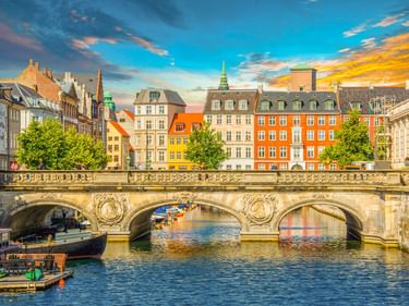 Steinbrücke mit drei Bögen über einem Kanal in Kopenhagens Altstadt. Bunte historische Gebäude säumen das Ufer unter dramatischem Abendhimmel.