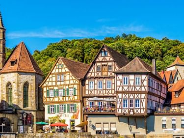Bunte Fachwerkhäuser und ein Kirchturm in der Altstadt von Schwäbisch Hall, mit grünen Hügeln im Hintergrund unter blauem Himmel.