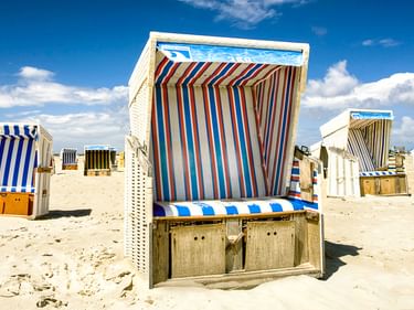 Bunte gestreifte Strandkörbe am sandigen Nordseestrand unter blauem Himmel. Weiße Korbstühle mit gestreiften Dächern in verschiedenen Farben.