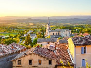 Luftaufnahme von Bonnieux mit Terrakotta-Dächern, einer Kirche mit Glockenturm und dem Luberon-Tal bei Sonnenuntergang in goldenem Licht.