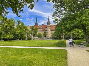 Schloss Merseburg mit Türmen und roten Dächern vom Schlossgarten aus gesehen. Ein Radfahrer geht auf einem Weg durch grüne Rasenflächen unter blauem Himmel.