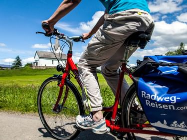 Radfahrer auf rotem Tourenrad mit blauen Radweg-Reisen Packtaschen auf Landstraße. Grüne Wiesen und weißes Haus unter blauem Himmel in Oberstaufen.
