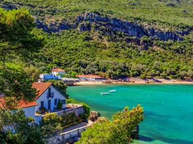 Weißes Haus mit rotem Dach mit Blick auf türkisfarbene Bucht im Parque Natural da Arrábida. Felsige Klippen mit grüner Vegetation erheben sich hinter dem Strand.