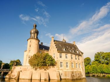 Burg Gemen in Borken, eine Wasserburg mit markanter Turmspitze und barocker Kuppel, umgeben von Wasser und grünen Bäumen unter blauem Himmel.