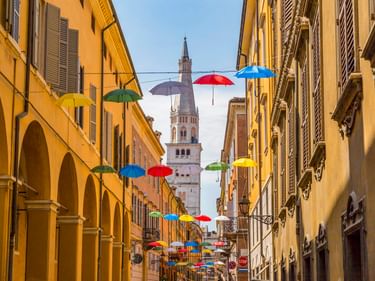 Enge Straße in Modena mit bunten Schirmen über der Gasse. Gelbe Gebäude mit Arkaden säumen die Seiten, ein Glockenturm ragt im Hintergrund auf.