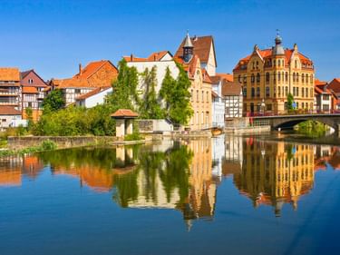 Bunte historische Gebäude mit orangen Ziegeldächern spiegeln sich in der ruhigen Weser in Eschwege. Eine Steinbrücke und ein Kirchturm unter blauem Himmel.