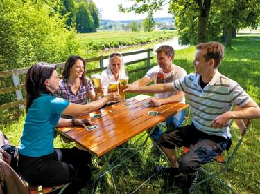 Fünf Freunde sitzen an einem Holztisch in einem Biergarten in Bayern und stoßen mit Biergläsern an. Grüne Wiesen und Bäume im Hintergrund.