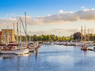 Eckernförder Hafen mit traditionellen Segelbooten und Yachten im ruhigen Wasser. Historische Gebäude und Bäume säumen die Uferpromenade unter blauem Himmel.