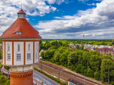 Luftaufnahme des Lingener Wasserturms mit markant rotem Kegeldach und weißem achteckigem Bau, umgeben von Bahngleisen und grüner Landschaft.