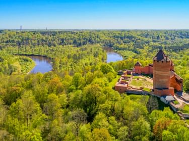 Luftaufnahme der roten Backsteinburg Turaida mit rundem Turm, umgeben von dichtem grünem Wald. Der Fluss Gauja schlängelt sich durch die Landschaft.