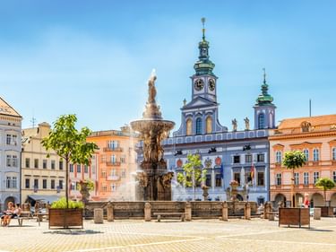 Samson-Brunnen auf einem sonnigen Platz in Budweis mit bunten Barockgebäuden und blauem Kirchturm mit Uhr im Hintergrund.