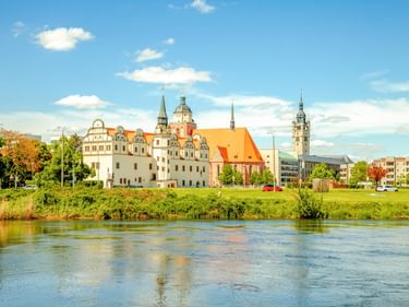 Residenzschloss Dessau mit weißer Fassade und Türmen an der Elbe, mit Kirche mit orangefarbenem Dach und grünem Flussufer unter blauem Himmel.