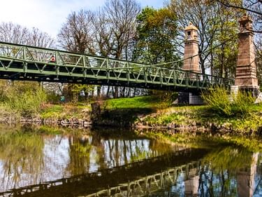 Grüne Metallbrücke über einen ruhigen Kanal mit zwei Steinpfeilern. Bäume säumen die Ufer, Spiegelungen im Wasser am Bodensee.