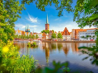 Historische Lübecker Obertrave-Uferpromenade mit bunten Gebäuden und Kirchtürmen, die sich im ruhigen Wasser spiegeln, eingerahmt von grünem Laub.