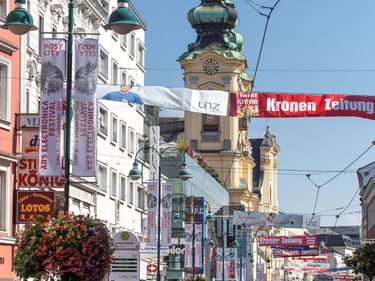Fußgängerstraße in Linz mit Straßenbahnschienen, Radfahrern und historischem Kirchturm mit grüner Kupferkuppel. Bunte Gebäude und Blumenampeln säumen die Straße.
