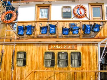 Holzdeck des Schiffes MS Panagiota mit blauen Radtaschen am Geländer. Orangefarbene Rettungsringe und Bullaugen am weißen Oberdeck.